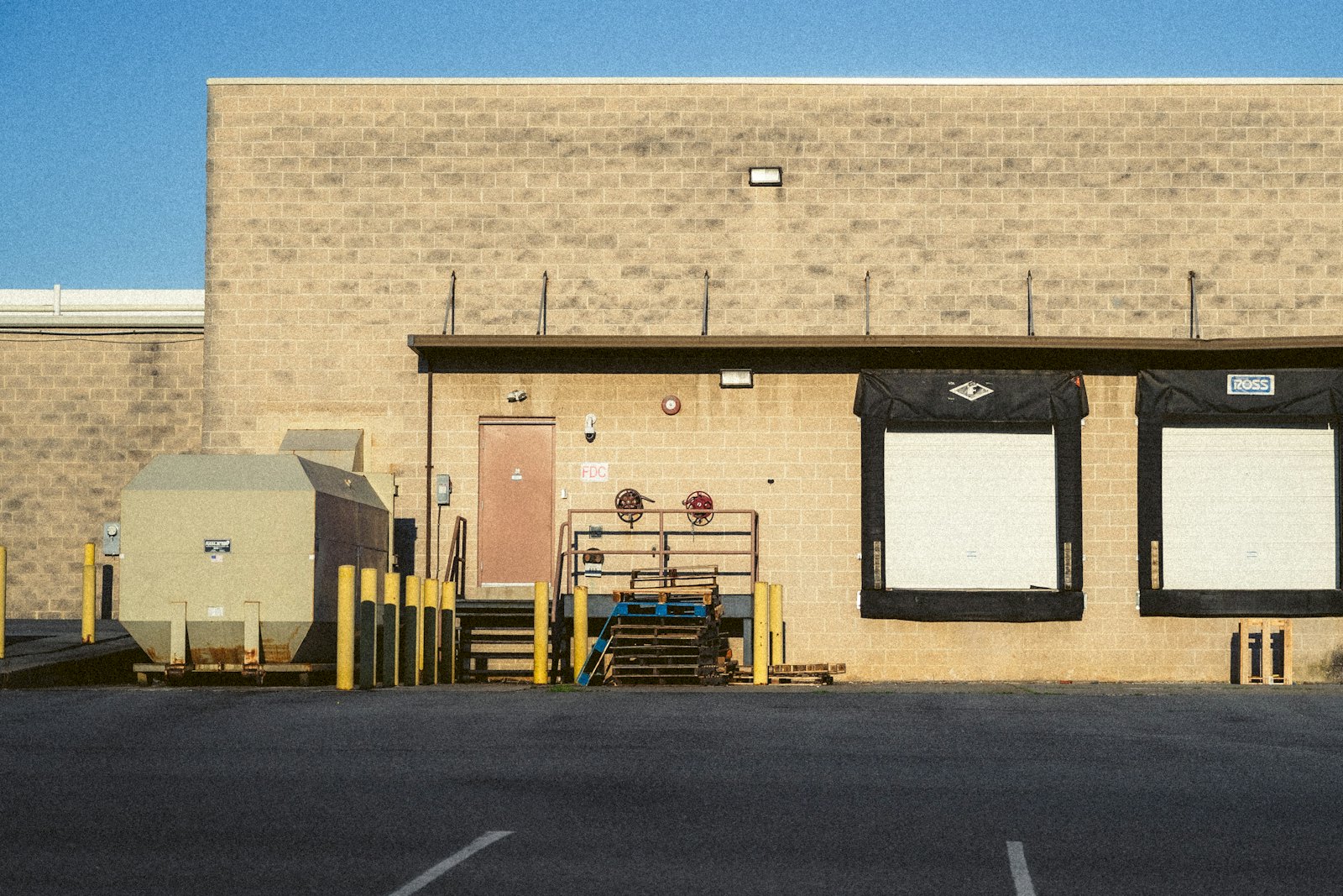 Late afternoon light on a tan block warehouse in an industrial park with two empty dock doors, black dock seals, yellow bollards and stacked wood pallets, the kind of back dock where RMA Fleet spots a rented storage trailer in the Cincinnati Tri-State