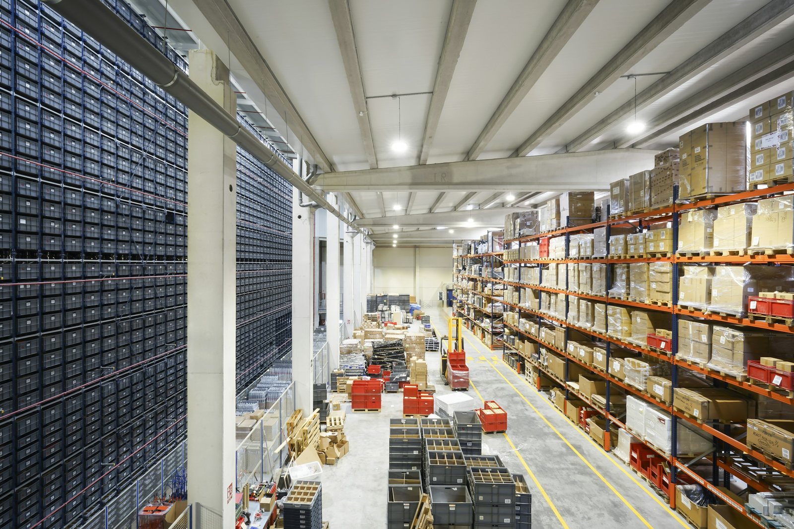Wide view down a warehouse aisle with shrink wrapped pallets in orange racking on one side and cartons, totes and pallets staged on the floor for shipment, the kind of partial load RMA Fleet picks up and delivers the same day in the Cincinnati Tri-State
